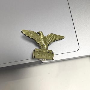 Könnte beinhalten: Bronzefarbene Metallbrosche mit einem detaillierten Adler mit ausgebreiteten Flügeln. Unter dem Adler befindet sich ein rechteckiges Banner mit den Buchstaben "SPQR". Der Anstecker ist wahrscheinlich ein historisches oder symbolisches Accessoire.