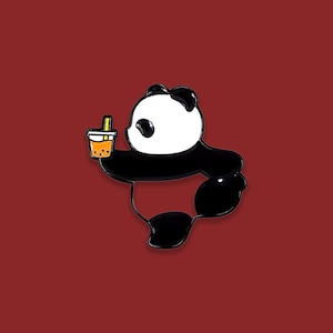 May include: A black and white panda bear enamel pin holding a cup of bubble tea with a yellow straw. The pin has a glossy finish and is set against a solid red background.