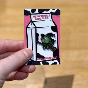 Op de afbeelding: Een groene hekskoe met een zwarte hoed en een bezem die uit een melkpak vliegt. Het pak heeft de tekst "Iedereen verdient een kans om te vliegen" en de tekst "@fuglycowdesigns".