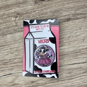 Può includere: Spilla raffigurante una mucca con un vestito rosa e una corona, all'interno di un cerchio. La spilla è ambientata su un cartone del latte rosa e nero con la scritta "WICKED" e il testo "IT'S GOOD TO SEE ME INST IT!" sul cartone.