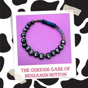 May include: A black beaded bracelet with the words "LIVE A LITTLE LIFE" in white letters. The bracelet has gold and blue beads. The image is on a purple background with the text "THE GURIOUS CASE OF BENJAMIN BUTTON" below.