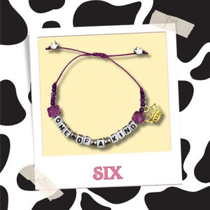 Puede incluir: Una pulsera de hilo morado con cuentas de letras blancas que deletrean "ONE OF A KIND". La pulsera presenta un dije de corona dorada y está acentuada con cuentas moradas y tapas de extremo plateadas en forma de corona. La palabra "SIX" está impresa en rosa.