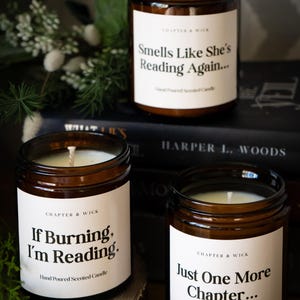 Op de afbeelding: Drie bruine glazen potkaarsen met witte etiketten en zwarte tekst. De etiketten luiden "Smells Like She's Reading Again...", "If Burning, I'm Reading." en "Just One More Chapter...". De kaarsen zijn handgegoten geurkaarsen.