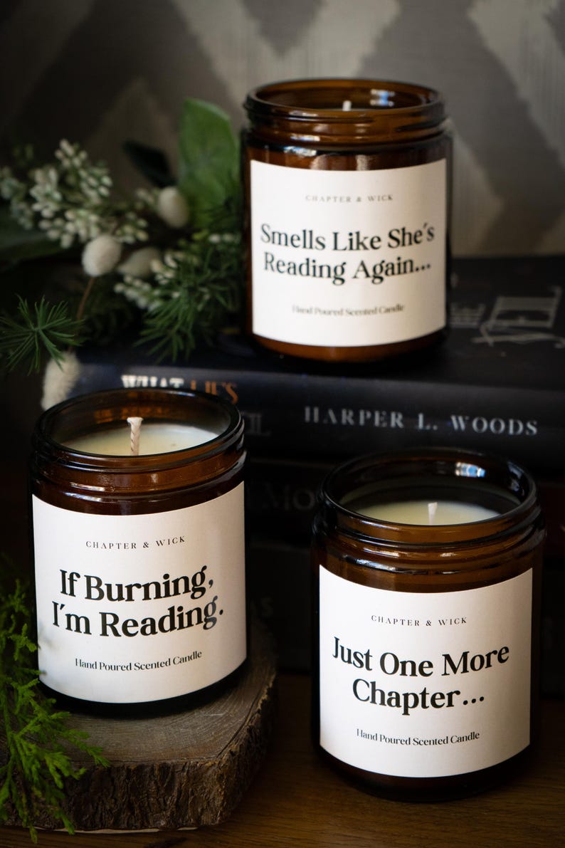 Op de afbeelding: Drie bruine glazen potkaarsen met witte etiketten en zwarte tekst. De etiketten luiden "Smells Like She's Reading Again...", "If Burning, I'm Reading." en "Just One More Chapter...". De kaarsen zijn handgegoten geurkaarsen.
