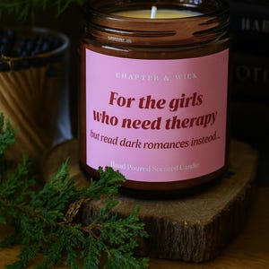 Puede incluir: Vela perfumada en un tarro de cristal marrón con una etiqueta rosa. La etiqueta dice: "Para las chicas que necesitan terapia pero leen romances oscuros en su lugar". La vela está sobre una rodaja de madera.