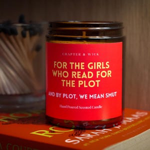 Puede incluir: Una vela con etiqueta roja con el texto "FOR THE GIRLS WHO READ FOR THE PLOT" está sobre un libro. La vela está en un tarro de cristal con tapa oscura. Un tarro de cristal con cerillas está en el fondo.