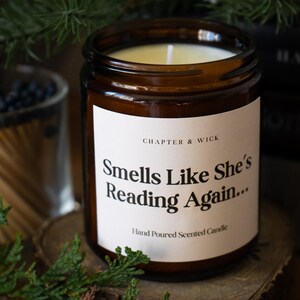 Op de afbeelding: Een bruine glazen kaarsenpot met een wit etiket met de tekst "Smells Like She's Reading Again..." en "Hand Poured Scented Candle". De kaars is omgeven door groen en boeken.