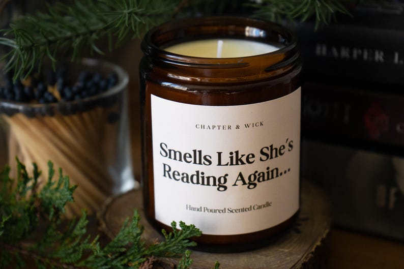 Op de afbeelding: Een bruine glazen kaarsenpot met een wit etiket met de tekst "Smells Like She's Reading Again..." en "Hand Poured Scented Candle". De kaars is omgeven door groen en boeken.