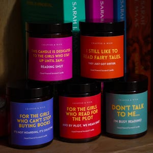 May include: Five scented candles in glass jars with black lids, each with a different colored label and text. The labels read: "This candle is dedicated to the girls who stay up until 3am... Reading Smut."