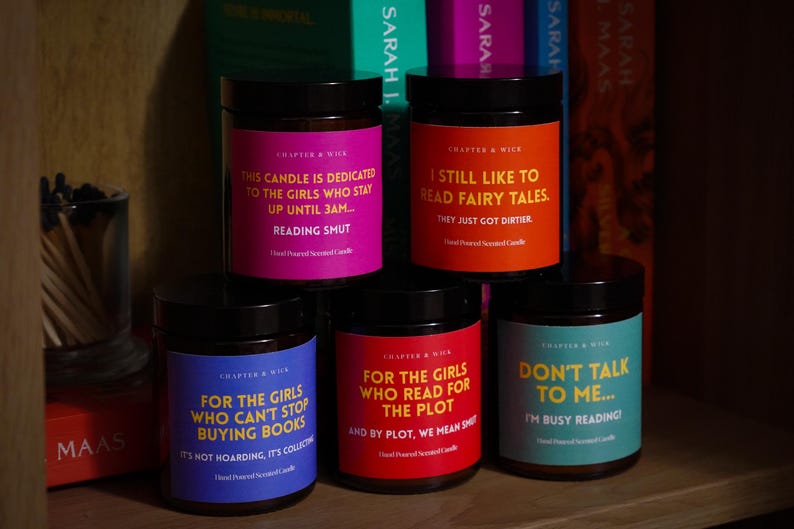 May include: Five scented candles in glass jars with black lids, each with a different colored label and text. The labels read: "This candle is dedicated to the girls who stay up until 3am... Reading Smut."