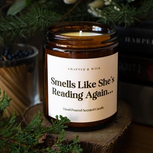 Op de afbeelding: Een bruine glazen potkaars met een wit etiket met de tekst "Smells Like She's Reading Again..." en "Hand Poured Scented Candle". De kaars staat op een houten schijf, met boeken en groen op de achtergrond.