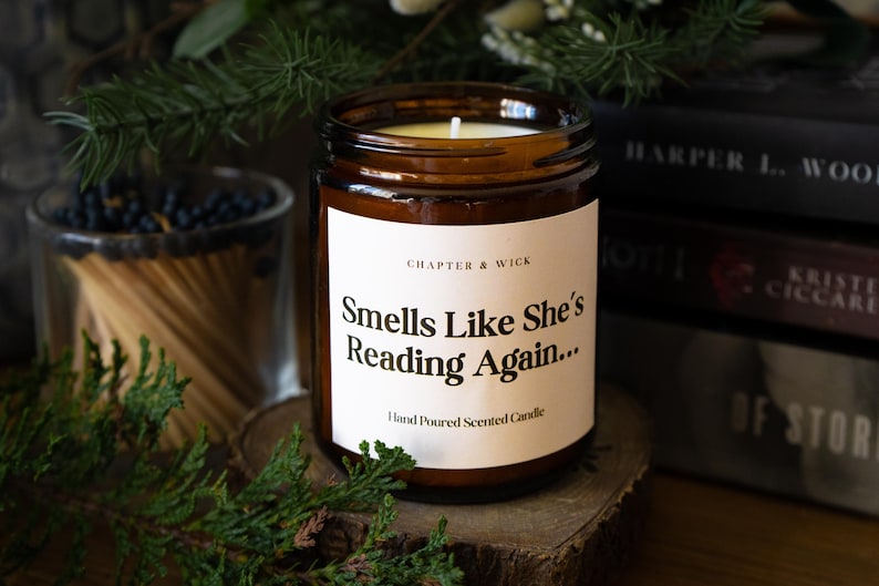 Op de afbeelding: Een bruine glazen potkaars met een wit etiket met de tekst "Smells Like She's Reading Again..." en "Hand Poured Scented Candle". De kaars staat op een houten schijf, met boeken en groen op de achtergrond.