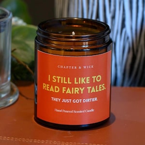 May include: A brown glass jar candle with an orange label that reads "I STILL LIKE TO READ FAIRY TALES. THEY JUST GOT DIRTIER." The candle is hand-poured and scented. The candle is on a brown surface.
