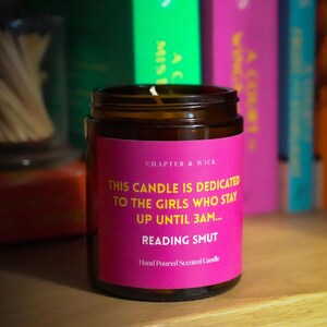 May include: A brown glass jar candle with a pink label that reads "THIS CANDLE IS DEDICATED TO THE GIRLS WHO STAY UP UNTIL 3AM... READING SMUT". The candle is on a wooden shelf with books in the background.