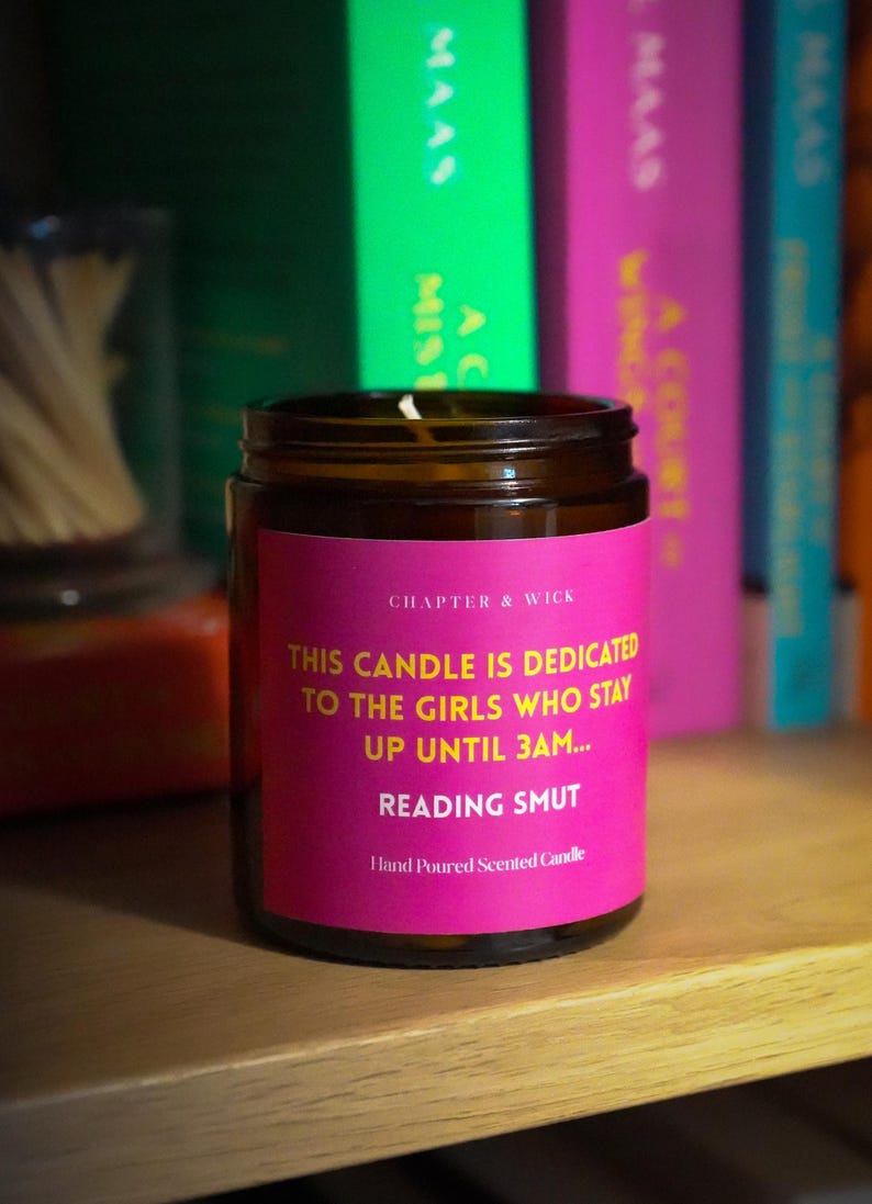 May include: A brown glass jar candle with a pink label that reads "THIS CANDLE IS DEDICATED TO THE GIRLS WHO STAY UP UNTIL 3AM... READING SMUT". The candle is on a wooden shelf with books in the background.