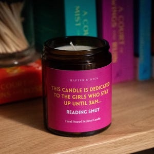 May include: A dark glass jar candle with a pink label. The label reads "THIS CANDLE IS DEDICATED TO THE GIRLS WHO STAY UP UNTIL 3AM... READING SMUT." The candle is on a wooden surface with books in the background.