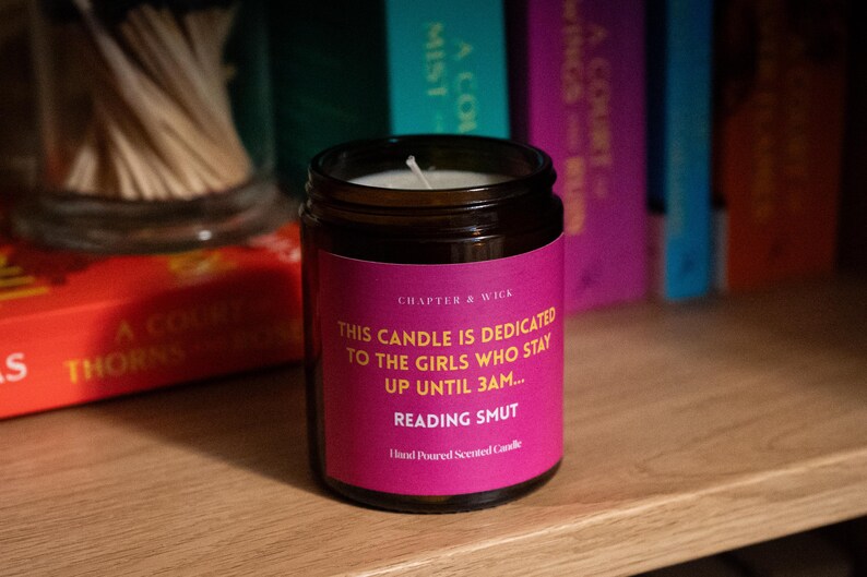 May include: A dark glass jar candle with a pink label. The label reads "THIS CANDLE IS DEDICATED TO THE GIRLS WHO STAY UP UNTIL 3AM... READING SMUT." The candle is on a wooden surface with books in the background.