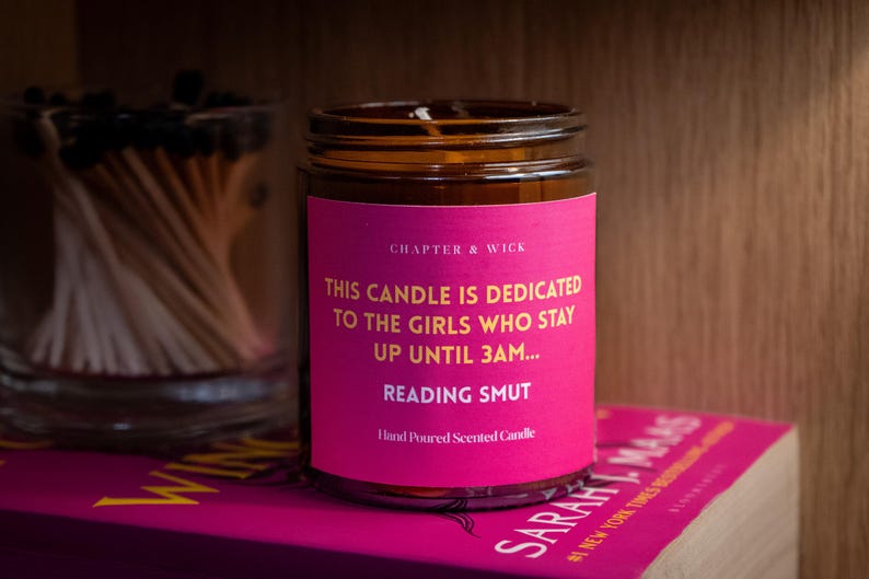 May include: A scented candle in a glass jar with a pink label that reads "THIS CANDLE IS DEDICATED TO THE GIRLS WHO STAY UP UNTIL 3AM... READING SMUT." The candle sits on a book and next to a glass jar of matches.