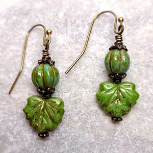 May include: A pair of green and brown dangle earrings with a leaf design. The earrings are made with glass beads and have a brass hook.
