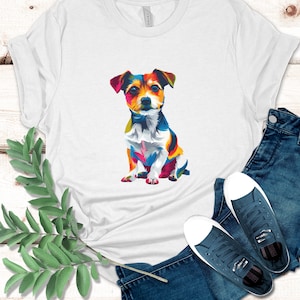 May include: White t-shirt featuring a colorful, pop art style illustration of a small dog. The dog has a multicolored coat with shades of red, blue, yellow, and white. The shirt is paired with blue jeans and navy blue sneakers.