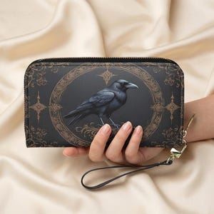 May include: Black wallet featuring a raven design. The wallet has a black raven inside a brown oval frame with decorative details. It has a zip closure and a wrist strap. The background is a light beige colour.