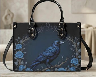 May include: Black tote bag featuring a blue raven perched on a branch, surrounded by blue roses and branches. The bag has black handles and a detachable shoulder strap. The design is set against a dark blue background.