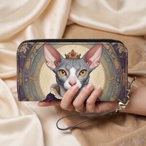 Könnte beinhalten: Eine Geldbörse mit Reißverschluss, die eine königliche Sphynx-Katze mit Krone zeigt. Die Katze ist im Porträtstil mit einem dekorativen Hintergrund aus Lila und Gold dargestellt. Die Geldbörse hat eine Handschlaufe und einen schwarzen Reißverschluss.