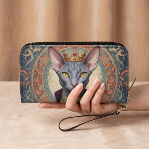 Könnte beinhalten: Schwarze Geldbörse mit Reißverschluss und einer Illustration einer königlichen Katze mit Krone und lila Umhang. Die Katze ist vor einem dekorativen blau-goldenen Hintergrund platziert. Die Geldbörse hat eine Handschlaufe.
