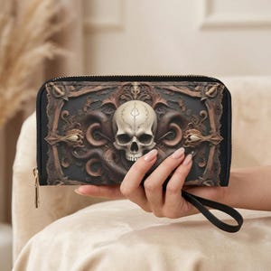 Könnte beinhalten: Schwarze Geldbörse mit Totenkopf-Design. Die Geldbörse hat einen detaillierten Totenkopf in der Mitte, umgeben von aufwendigen, dunkelbraunen Verzierungen. Sie hat einen schwarzen Reißverschluss und eine Handschlaufe.