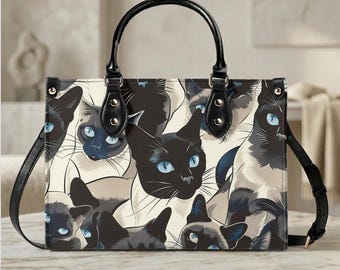 May include: A rectangular handbag featuring a repeating pattern of Siamese cats with blue eyes. The bag has black handles and a detachable shoulder strap. The cats are depicted in various poses against a cream background.