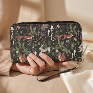 May include: Black zip-around wallet with a woodland pattern featuring squirrels, foxes, mushrooms, and foliage. The wallet has a wrist strap and gold-tone hardware. The design is printed on the front and back of the wallet.