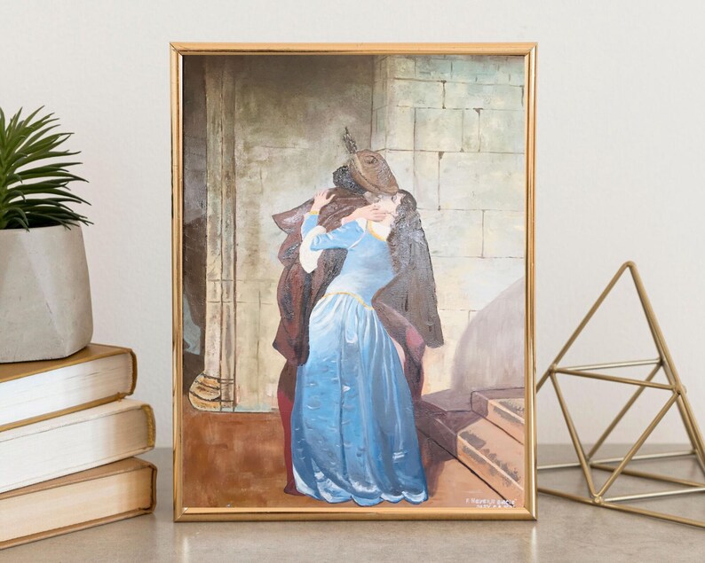 Puede incluir: Una pintura enmarcada representa a una pareja abraz&aacute;ndose, ambientada en una arquitectura de piedra. La mujer viste un largo vestido azul fluido, mientras que el hombre est&aacute; envuelto en ropa oscura. La obra de arte est&aacute; enmarcada en oro.