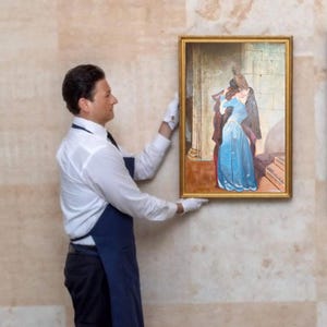 Puede incluir: Un cuadro enmarcado que representa a una pareja abraz&aacute;ndose, la mujer con un vestido azul. El cuadro lo sostiene una persona con camisa blanca, pantal&oacute;n negro, delantal azul y guantes blancos. El marco es dorado.