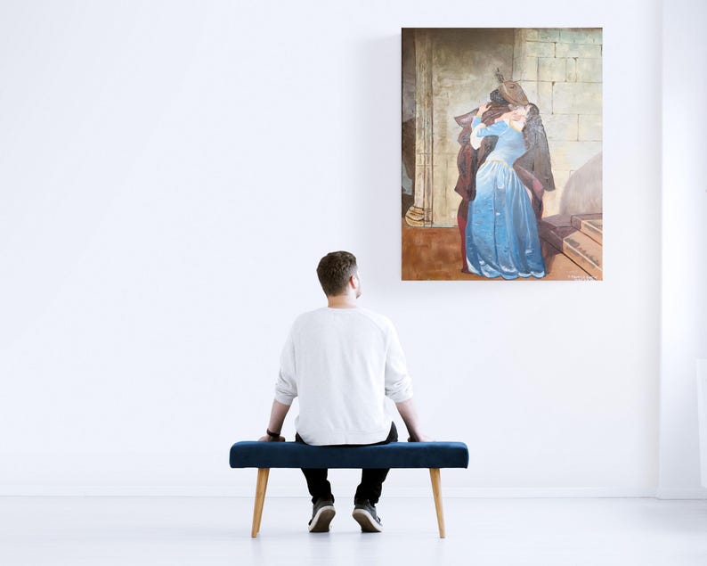 Puede incluir: Una pintura enmarcada que representa a una mujer con un vestido azul y un hombre con un abrigo oscuro. La obra de arte se exhibe en una pared blanca sobre un banco azul. Una persona est&aacute; sentada en el banco, mirando la pintura.