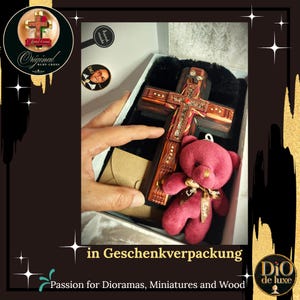 K&ouml;nnte beinhalten: Ein handgefertigtes Holzkreuz mit aufwendigen Intarsien, pr&auml;sentiert in einer Geschenkbox mit einem roten Teddyb&auml;ren. Das Kreuz hat eine dunkle Holzmaserung und dekorative Akzente. Der Text "Original Ruby-Cross" ist sichtbar.
