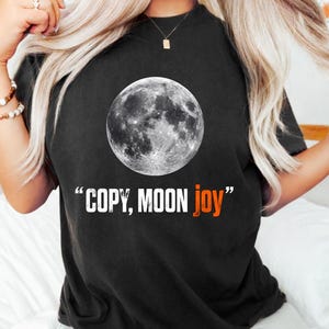 Può includere: T-shirt nera con un'immagine dettagliata in scala di grigi della luna. Sotto la grafica della luna, il testo recita "COPY, MOON joy" in bianco e arancione. La maglietta ha un girocollo e maniche corte.