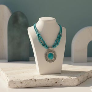 May include: A turquoise beaded necklace with a large pendant on a white display. The pendant has a silver-coloured frame with a turquoise stone centre. The necklace is displayed on a white bust against a neutral background.