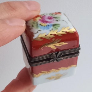 May include: A small, square porcelain trinket box with a red base and a white lid. The lid is decorated with hand-painted flowers in shades of pink, yellow, blue, and green. The box has a gold trim and a hinged lid with a clover-shaped clasp.