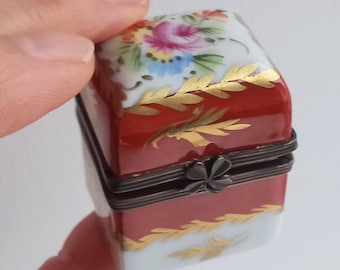 Puede incluir: Una pequeña caja de porcelana cuadrada con una base roja y una tapa blanca. La tapa está decorada con flores pintadas a mano en tonos de rosa, amarillo, azul y verde. La caja tiene un borde dorado y una tapa con bisagras con un cierre en forma de trébol.