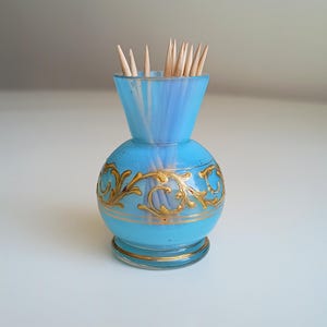 Puede incluir: Un jarrón de vidrio azul claro con diseños florales dorados, lleno de palillos de dientes de madera.