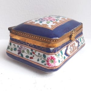 May include: An antique porcelain trinket box. The lid is navy blue with a floral design and gold trim. The sides are white with floral and leaf patterns. The box has a gold clasp.
