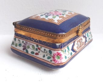 Puede incluir: Una caja de baratijas rectangular de porcelana antigua. La tapa es azul marino con un diseño floral y ribete dorado. Los lados son blancos con patrones florales y de hojas. La caja tiene un cierre dorado.