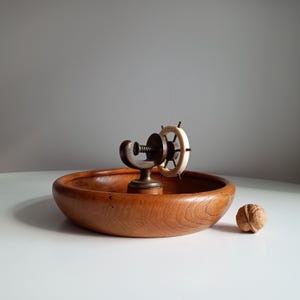 May include: A wooden bowl with a brass nutcracker in the center, featuring a small ship's wheel design. A single walnut sits to the right of the bowl. The bowl and nutcracker have a warm, brown color, and the wheel is white.