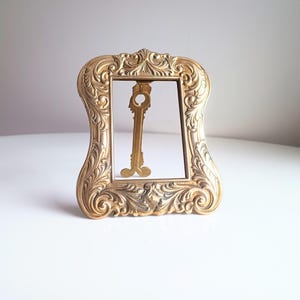 May include: Ornate gold-colored picture frame with a key-shaped object inside. The frame has a detailed, scrolling design. The key is also gold and has a decorative head and bow.