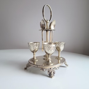 May include: A silver-toned egg cup stand with four egg cups and spoons. The egg cups have a decorative pierced design. The stand has a round base with ornate feet and a handle for carrying. A classic and elegant serving piece.