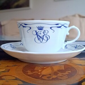 Op de afbeelding: Witte theekop en schotel set met blauwe details. De theekop heeft een blauw kroon- en monogramontwerp. De schotel heeft een bijpassend blauw patroon langs de rand. De set staat op een houten oppervlak.