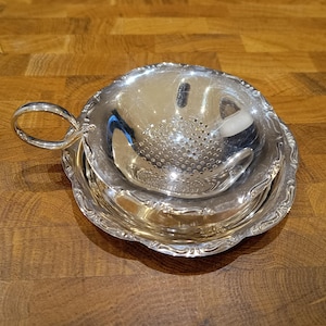Op de afbeelding: Een zilveren thee-ei met een handvat, rustend op een bijpassende schotel. Het thee-ei heeft een geperforeerde kom voor het trekken van theebladeren. De set is gemaakt van glanzend, reflecterend metaal en ligt op een houten oppervlak.