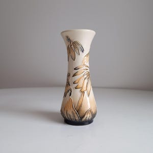 Puede incluir: Un pequeño jarrón de cerámica de color crema con un diseño floral. El jarrón tiene un cuello estrecho y una base ancha, y está decorado con flores marrones y amarillas.