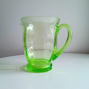 Può includere: Una brocca vintage in vetro verde con manico e base decorativa. La brocca ha un corpo trasparente e un manico e una base verde brillante. Il vetro ha un design a coste sui lati. La brocca viene probabilmente utilizzata per servire bevande.
