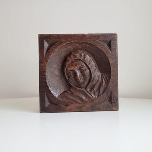 Puede incluir: Talla de madera cuadrada de color marrón oscuro que representa a una persona con una prenda con capucha. El relieve está dentro de un marco circular, sobre un fondo cuadrado con detalles en las esquinas. La veta de la madera es visible.